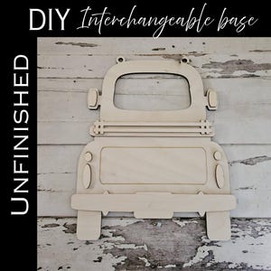 May include: Unfinished wooden truck-shaped base for DIY projects. The truck has a cab, bed, and wheels, with cutouts for interchangeable decorations. The word "UNFINISHED" is printed vertically on the left, and "DIY Interchangeable base" is written at the top.