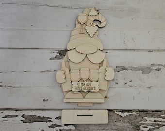 May include: Wooden gnome figurine holding a sign that says "At my age I need glasses". The gnome is wearing a hat with a grape design and has a beard. The gnome is holding four wine glasses in front of him.