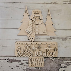 Könnte beinhalten: Ein heller Holzausschnitt, der einen Schneemann mit einem Hut mit der Aufschrift FROSTY, einem Schal und drei Knöpfen zeigt, flankiert von zwei Tannenbäumen. Darunter befindet sich ein rechteckiges Schild mit dem Text hello winter und dem Wort SNOW.