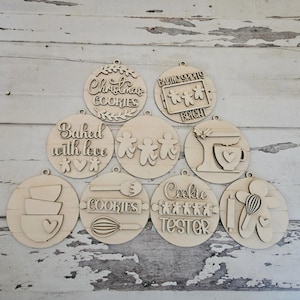 May include: A collection of wooden Christmas ornaments with baking-themed designs. Each ornament is round and features laser-cut text and images, including phrases like "Christmas Cookies" and "Baking Spirits Bright."