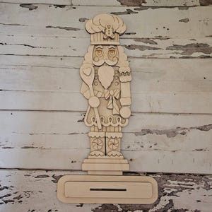 May include: A light-coloured wooden nutcracker cutout featuring a chef hat with a small gingerbread man detail, a moustache, and a heart-shaped spoon. The figure stands on a rectangular base with a horizontal slot, showcasing intricate carved details.