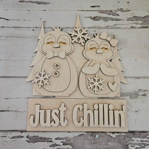 Könnte beinhalten: Ein helles Holzschild mit zwei ausgeschnittenen Pinguinen mit geschlossenen Augen, Fliege, Haarschleife und Schneeflocken-Akzenten. Die Pinguine stehen vor stilisierten Tannenbäumen. Darunter befindet sich eine rechteckige Holzplatte mit dem Schriftzug Just Chillin' in erhabenen, dunkel umrandeten Holzbuchstaben.