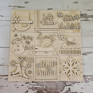 Può includere: Nove quadrati di legno con disegni a tema invernale. I quadrati presentano un pupazzo di neve, un cappello, fiocchi di neve, pattini da ghiaccio e le parole "hello winter", "winter wonderland", "baby it's cold", "frosty", "let it snow" e "brrrr".