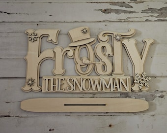 May include: Wooden cutout of the words "Frosty the Snowman" with a snowman head and hat in the center. The cutout is layered and has a stand.