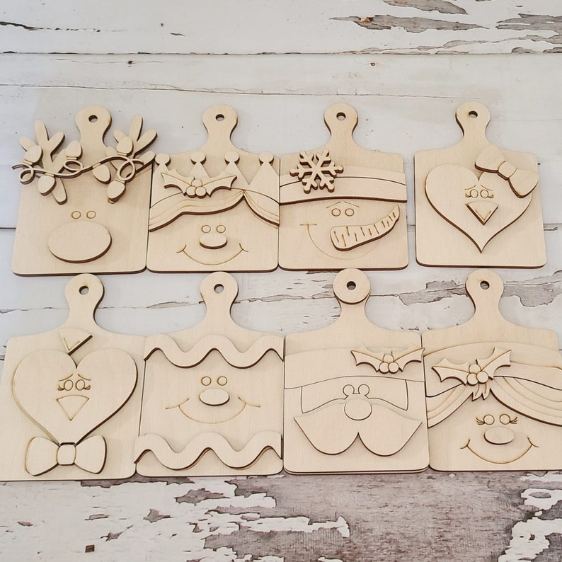 K&ouml;nnte beinhalten: Acht unfertige Holzschneidebretter mit festlichen Weihnachtsdesigns. Jedes Brett zeigt ein anderes Gesicht, darunter ein Rentier, ein Schneemann und der Weihnachtsmann. Die Bretter sind rechteckig mit einem Griff und aus hellfarbigem Holz.