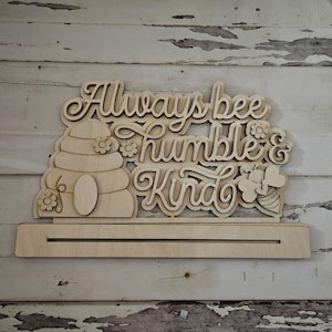May include: Wooden sign with the text "Always bee humble & kind" and a beehive design. The sign is on a stand.