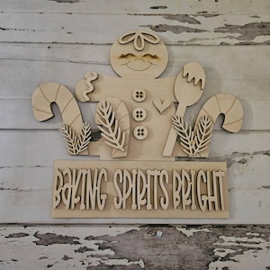 May include: A light-colored wooden cutout of a gingerbread person holding a spoon, flanked by candy canes and pine sprigs. The figure features three circular buttons and a heart detail. Below, a rectangular sign displays the text BAKING SPIRITS BRIGHT.