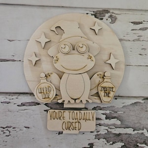 May include: A circular light-colored wooden cutout featuring a frog wearing a hat, surrounded by four-pointed stars. The frog holds two bottles labeled Lily Pad Lixir and Toadstool Tonic. A rectangular sign below reads Youre Toadally Cursed.
