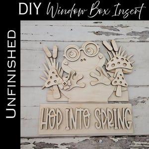 May include: Unfinished wooden DIY window box insert featuring a frog wearing glasses, mushrooms, and the phrase "Hop Into Spring." The frog has outstretched hands and is surrounded by decorative elements. The word "UNFINISHED" is printed vertically on the left.