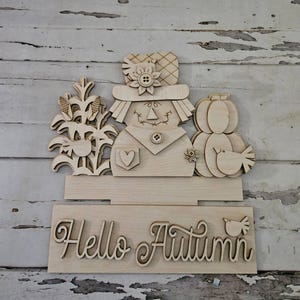 Könnte beinhalten: Holzschild zum Thema Herbst mit einer Vogelscheuche, einem Kürbis und Maiskolben. Das Schild enthält den Satz "Hello Autumn" in Schreibschrift sowie einen kleinen Vogelausschnitt. Das Design ist aus Naturholz gefertigt.