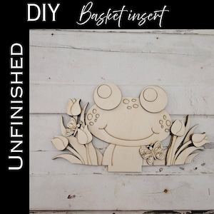 May include: Unfinished wooden frog basket insert with tulip and butterfly accents. The DIY craft piece features the text "DIY Basket insert" and "UNFINISHED". The frog has large eyes and a smiling face, perfect for spring decor.