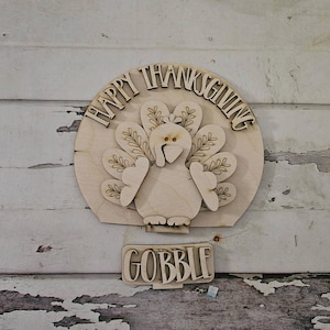 May include: A wooden Thanksgiving decoration featuring a turkey with a fan of feathers, the words "HAPPY THANKSGIVING" in a semi-circle above the turkey, and the word "GOBBLE" below. The turkey and text are cut from light-colored wood.