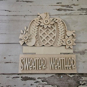 May include: Wooden fall-themed sign with a pumpkin design, sunflowers, and birds. The sign includes the words "SWEATER WEATHER" cut out at the bottom. The sign is made of unfinished wood.