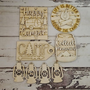May include: Wooden cutouts for crafting, featuring camping themes.  The cutouts include a sign that says "Happy Camper", a sign that says "Camp", a sign that says "Life is Better Around the Campfire", a mason jar with the text "Collect Memories", and four lantern shapes.