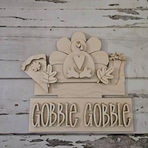 May include: A wooden Thanksgiving decoration featuring a turkey with a heart-shaped chest, surrounded by leaves and a flower. The words "GOBBLE GOBBLE" are displayed below the turkey. The decoration is made of light-colored wood.