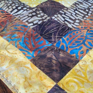 May include: A colorful, patterned table runner with a patchwork design. The runner features diamond shapes in shades of yellow, brown, purple, and blue. The fabric has a batik-like appearance, adding visual interest to the home decor item.
