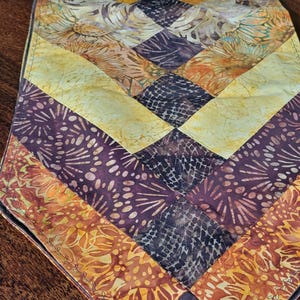 May include: A quilted table runner with a geometric pattern. The runner features a series of diamond shapes in shades of yellow, orange, brown, and black. The fabric has a floral and abstract design.