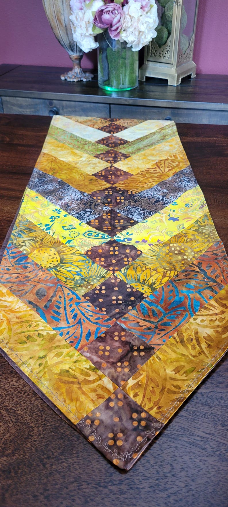 May include: A vibrant, quilted table runner with a chevron pattern. It showcases a mix of warm hues like yellow, brown, and orange, with floral and geometric designs. The edges are finished with decorative stitching.