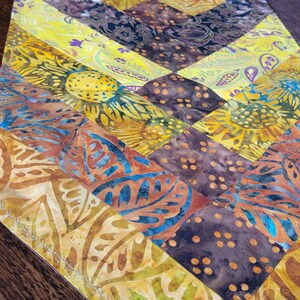 May include: A vibrant table runner with a patchwork design. The runner features a variety of patterns and colours, including yellow, brown, and blue. The design includes floral and geometric shapes.