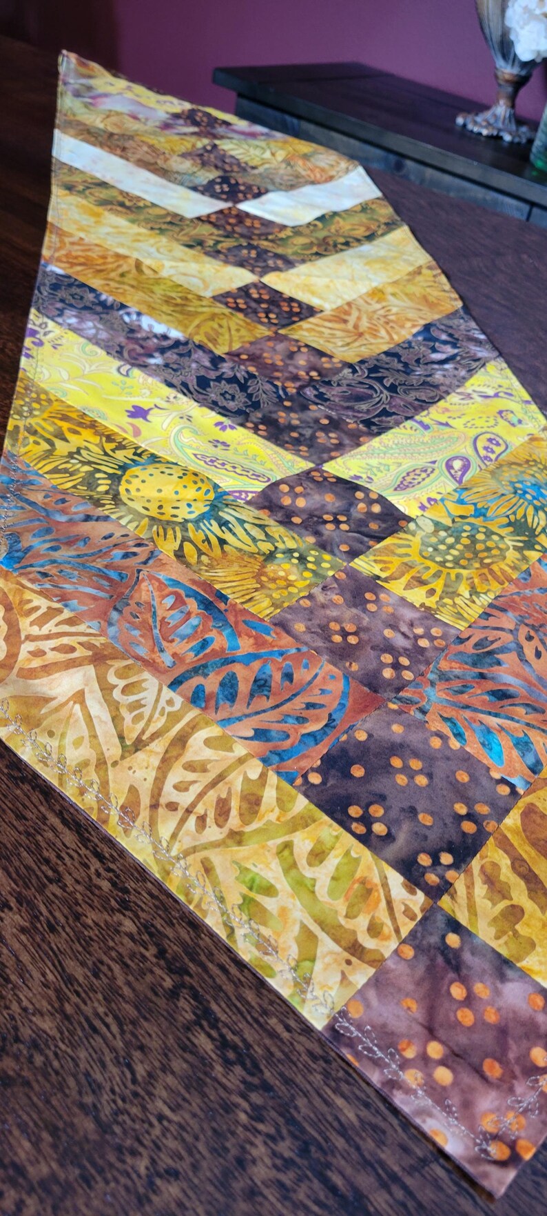 May include: A vibrant table runner with a patchwork design. The runner features a variety of patterns and colours, including yellow, brown, and blue. The design includes floral and geometric shapes.