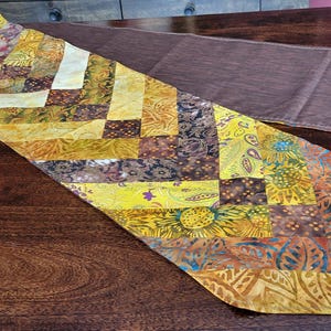 May include: A patchwork table runner in shades of yellow, brown, and orange, with a second plain brown runner in the background. The patterned runner is placed on a dark wooden surface. The patchwork design features geometric patterns.