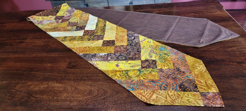 May include: A patchwork table runner in shades of yellow, brown, and orange, with a second plain brown runner in the background. The patterned runner is placed on a dark wooden surface. The patchwork design features geometric patterns.