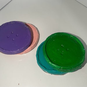 Puede incluir: Dos posavasos redondos y coloridos con un diseño de botón. El posavasos superior es morado con una base rosa. El posavasos inferior es verde con una base turquesa.