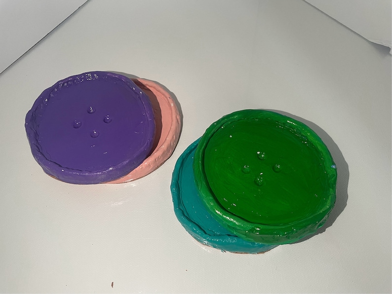 May include: Two round, colorful coasters with a button-like design. The top coaster is purple with a pink base. The bottom coaster is green with a teal base.