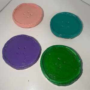 May include: Four round coasters with a button design. The coasters are painted in pink, teal, purple, and green.