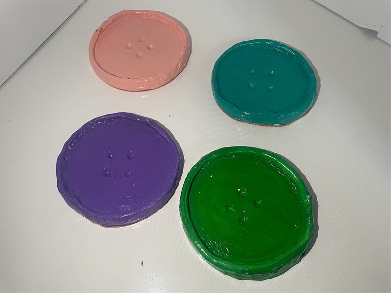 May include: Four round coasters with a button design. The coasters are painted in pink, teal, purple, and green.