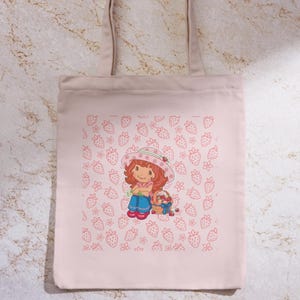 Könnte beinhalten: Hellrosa Tragetasche mit einer Cartoon-Illustration eines Mädchens mit roten Haaren, Hut und blauer Hose, das einen Korb mit Erdbeeren hält. Der Hintergrund hat ein Muster aus Erdbeeren und Blumen.