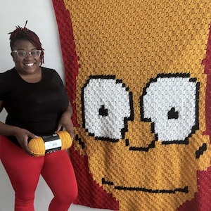 May include: A person is holding a ball of yellow yarn in front of a large crochet blanket featuring a cartoon character with yellow hair, white eyes, and a black mouth. The blanket is made with red, yellow, white, and black yarn.