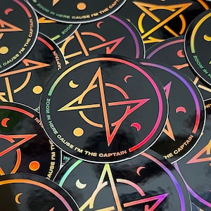 May include: A close-up of a pile of round stickers with a black background and a rainbow-colored design. The stickers feature a pentagram with a circle around it and the text "Zoom in here cause I'm the captain".