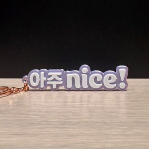Puede incluir: Un llavero morado con una inscripción blanca que dice "야주 nice!" El llavero está hecho de un material suave y flexible y tiene un anillo de metal plateado.