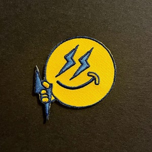 以下が含まれることがあります： スマイリーフェイスと2つの稲妻が描かれた黄色い刺繍パッチ。スマイリーフェイスの側面から青い稲妻が出ています。