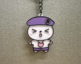 Pode incluir: Um chaveiro com um personagem de desenho animado branco com um boné roxo, um coração rosa e a letra "A" no boné. O personagem tem uma expressão de surpresa com a boca aberta.