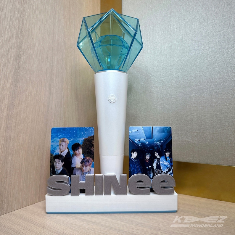 K&ouml;nnte beinhalten: Ein wei&szlig;er Lichtstick mit einer blauen, diamantf&ouml;rmigen Spitze, zwei Foto-Karten einer Boyband und das Wort "SHINee" in silbernen Buchstaben auf einem wei&szlig;en St&auml;nder.