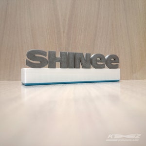 K&ouml;nnte beinhalten: Ein 3D-gedrucktes Namensschild mit dem Wort "SHINee" in silbergrauen Buchstaben auf einem wei&szlig;en Sockel mit einer hellblauen Umrandung.