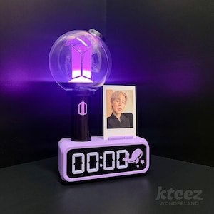 Puede incluir: Un bastón de luz morado y negro con una parte superior esférica transparente, que muestra un logotipo morado. Se encuentra encima de un reloj lavanda con una pantalla digital que muestra "00:00". Una foto de una persona se coloca junto al bastón de luz. La imagen tiene el texto "kteez WONDERLAND".