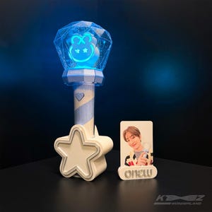 Puede incluir: Un palo de luz azul con un diseño de oso y una base en forma de estrella. El palo de luz tiene un brillo azul y la palabra "onew" es visible. También hay una tarjeta fotográfica con la palabra "onew".
