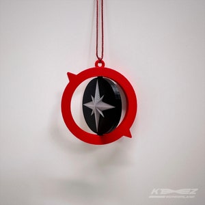 May include: A red and black ornament with a spinning compass design. The compass is black with a silver star in the center. The ornament is hanging from a red string.