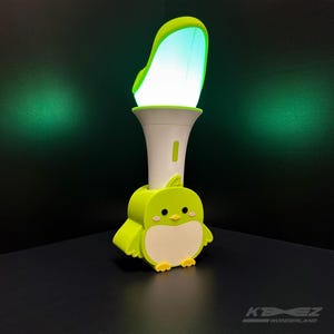 Può includere: Una luce a forma di uccello dei cartoni animati verde e bianca. La luce ha una base bianca, un corpo verde a forma di uccello e una parte superiore curva e luminosa. La luce è su una superficie nera.