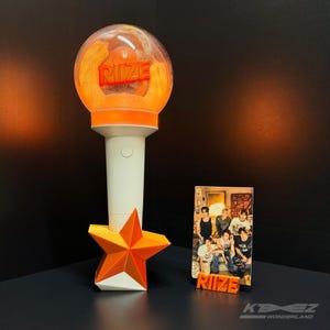May include: An orange and white light stick with the word "RIZE" in orange letters. The light stick has a star-shaped base. A photo of a group of people is next to the light stick, with the word "RIZE" in orange letters.