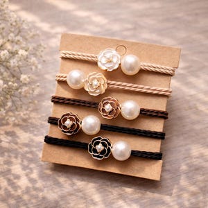 May include: A set of six hair ties on a cardboard backing. Each tie features a different color of cord: beige, light brown, dark brown, and black. Each tie has a flower-shaped accent with a pearl center and a large pearl bead.