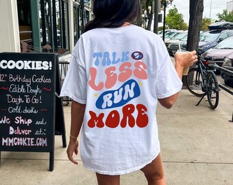 May include: White t-shirt with a graphic design that says "Talk Less Run More" in a retro font. The graphic is in shades of pink, blue, and red.