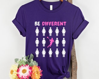 May include: Purple t-shirt with the text "BE DIFFERENT" and a graphic of a lacrosse player in pink, surrounded by white stick figures.