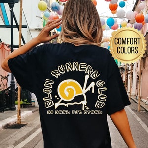 May include: Black t-shirt with a yellow and white snail graphic and the text "SLOW RUNNERS CLUB, NO NEED FOR SPEED." The shirt also has the text "COMFORT COLORS" and "BLACK" in the image.