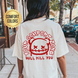 May include: Ivory-colored t-shirt with a red graphic on the back that reads "Comfort Zone Will Kill You" around a cartoon face. The shirt is part of the "Comfort Colors" collection.