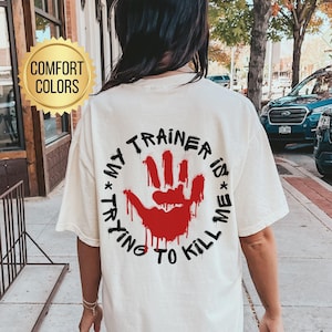 Puede incluir: Una camiseta blanca con un gráfico de huella de mano roja y el texto "My Trainer is Trying to Kill Me". La camiseta es de color confort y está disponible en marfil.