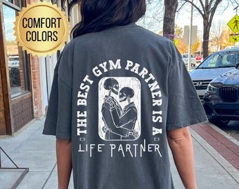 Op de afbeelding: Een donkergrijs oversized T-shirt met een witte grafische print van twee skeletten die elkaar omhelzen. De tekst op het shirt luidt "The best gym partner is a life partner".